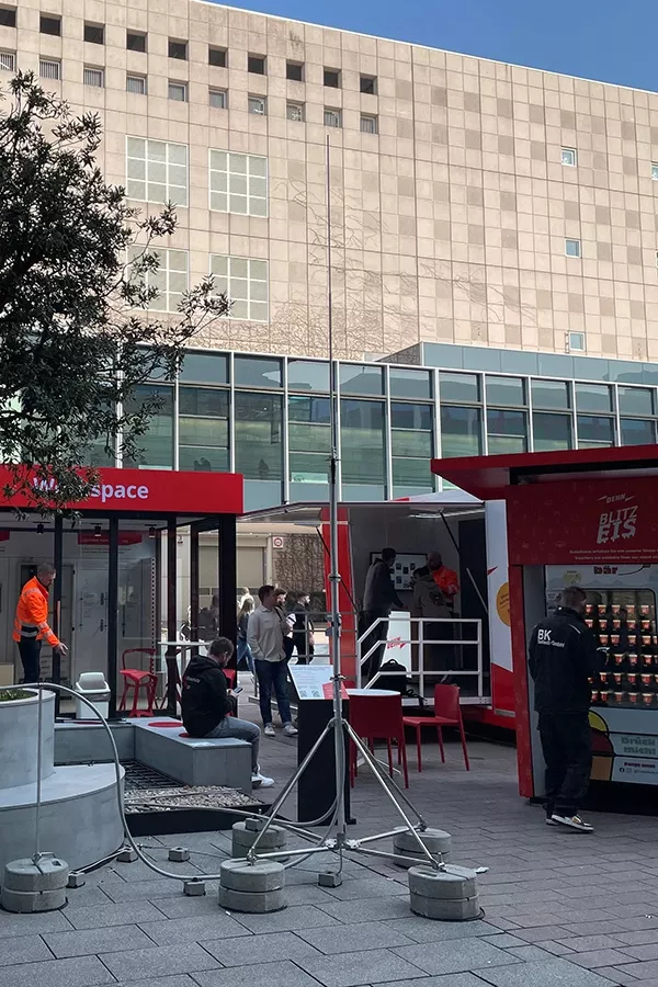 An outdoor urban area featuring a mobile food truck, a shipping container repurposed as a workspace, and several people engaged in various activities. The background includes a modern building with large windows.